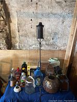 Photo showing assortment of glass and metal lanterns, tiki torch, lamps oil bottles, and wind chimes arranged on a table against wooden and plaster wall.