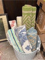 Six rolled runners of various patterns and colors bundled in a white basket among boxes and a framed picture.