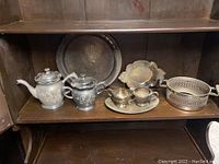 Full lot with all 10 pieces of silver plated serving ware on wooden shelf, showing floral engraved teapot, coffee pot, creamers, sugar bowl, oval tray, pierced basket, and large round tray.