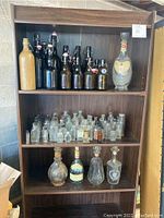 Wood shelf with amber and dark glass flip-top beer bottles with ceramic stoppers on top shelf. Clear glass vintage apothecary bottles on middle shelf. Decorative vintage whiskey decanters on bottom shelf.