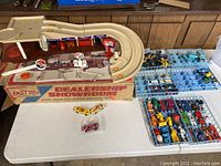 Overview of the assembled Kenner Fast 111's Dealership Showroom playset on a table with die-cast cars and trucks arranged in blue trays and three vehicles in front.