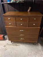 Full view of 5 drawer chest dresser with shell-shaped drawer pulls, standing on short legs on cement floor.