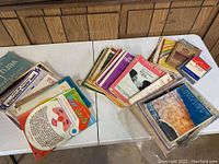 Photo of assorted music books and sheet music piled on a table, showing various colorful covers and titles including Christmas-themed sheet music.