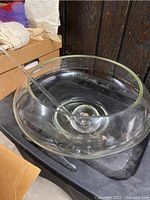 Clear glass punch bowl with wide opening and thick base on black surface, accompanied by clear glass ladle resting inside the bowl.