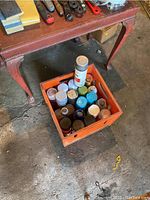 Photo showing orange crate containing multiple cans of spray paint in assorted colors under a table.