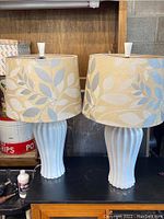Two light blue ceramic table lamps with beige shades featuring blue and white leafy pattern, placed on black surface.