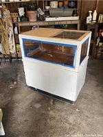 Full view of the vintage display case showing white painted wooden structure, glass top and glass sides, standing on a garage floor.