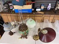 View of vintage oil-style lamp with glass chimney, green and white spherical glass shade, brass bracket, 4 hurricane glass globes, and other lamp parts on table.