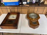 Two wall hanging clocks displayed on a table, one rectangular with a white dial, one octagonal with a silver dial, each with pendulums visible.