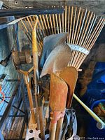 Tools arranged in metal stand including rakes, shovels, and axes showing wood handles and metal heads with signs of rust and wear.