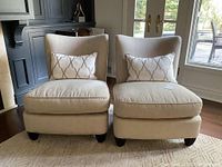 Front view of two cream upholstered armless chairs with matching lumbar pillows on rug in front of glass doors