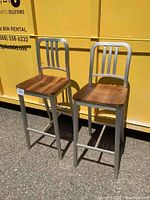 Front three-quarter view of two bar stools outdoors against a yellow bin on pavement