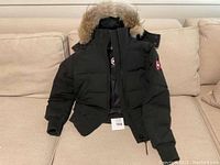 Front view of black Canada Goose bomber jacket on couch, showing zip closure, patch and fur hood