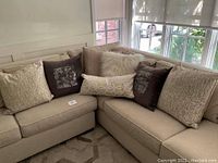 Wide view of eight decorative pillows arranged on a beige sectional sofa near large windows showing varied textures and colors.
