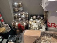 Box containing assorted large silver and gold Christmas ball ornaments, some still in plastic packaging.