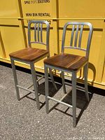 Two bar stools at a slight angle showing metal frame and wooden seats against a yellow backdrop