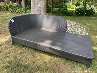 Full view of dark brown resin wicker chaise lounge on grass, showing curved backrest and flat seating area.
