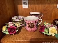 Photo showing two vintage moustache cups, three floral porcelain candle holders, and a Noritake Nippon china box arranged on wooden shelf.
