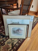 Three framed paintings stacked on the floor: one larger artwork behind two smaller ones, showing architectural scenes mainly of buildings.
