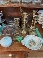 Group of brass candle sticks, Belleek ceramic container with lid, floral porcelain dish, and glass bowls on wooden shelf.