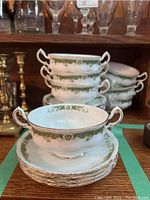 Stacked six consommé bowls and six saucers showing green floral and gold pattern with double handles on bowls.