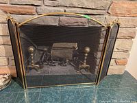 Front view of brass fireplace screen in front of stone fireplace, showing mesh panels and andirons behind screen.