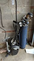 Golf bag filled with golf clubs, standing next to collapsible hand/pull cart, bike pump, and bicycle basket in a garage or storage area with concrete walls.
