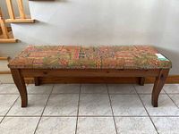 Full front view of wooden hall bench on tiled floor in front of wall and stairs, showing patterned upholstered seat and wooden legs.