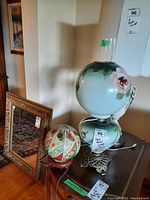 Two hand painted lamps on a table with a framed mirror nearby; larger floral lamp with glass chimney and smaller colorful glass globe lamp visible