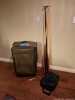 Full lot with large brown suitcase, seven wooden pool cues standing upright, and small black soft-sided bag on floor.