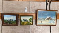 Three framed signed paintings displayed together on the floor. Each painting depicts a different landscape scene: a rural house on a hill, a country road with houses, and a winter barn scene.