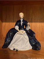 Front view of seated female porcelain figurine in a dark blue and white dress holding a book on her lap.
