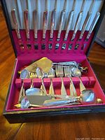 Open flatware case showing neatly arranged forks, spoons and serving pieces against a pink lined interior.