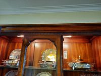 Three silver plated serving pieces displayed inside a wooden cabinet, showing overall style and grouping.