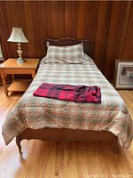 Full view of wooden single bed frame with mattress set up, plaid bedding, and nearby wooden side table with lamp.