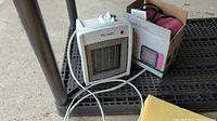 Two space heaters on a metal table, one white Pelonis heater approximately 8 inches tall with control knob and grill, the other SAi heater partially visible in box behind