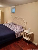 Bedroom view showing one white side table placed next to bed with floral sheets and iron headboard.