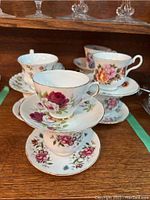 Eleven mixed floral tea cups and saucers stacked and displayed showing variety of rose and floral patterns with gold rims.