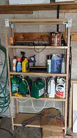 Full view of metal shelf holding assorted gardening and cleaning chemicals, tools, and accessories against a wood and concrete wall.