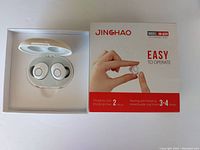 Open white charging case with pair of small white wireless hearing aids inside. Box for Jinghao model JH-A39 to the right with promotional details.