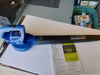 Blue electric leaf blower with long black nozzle shown on a white surface, photographed from above at an angle.