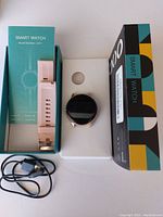 Smart watch displayed with its packaging; two boxes, the watch itself with black round face, pink band, and a USB charging cable.