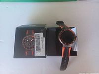 Photo of a black smartwatch with orange accents next to its box illustrating the watch image on the packaging. The watch has a round digital face and a black rubber strap with orange lining.