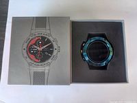 Photo showing smart watch in original box with box lid partially ajar displaying watch and packaging.