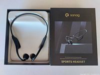 Photo of black Sanag sports headset inside its retail box alongside the product packaging box showing branding and image.
