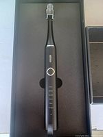 Photo showing the Oral-B electric toothbrush inside its original packaging, black toothbrush with mode indicators and brush head cover attached.