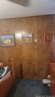 Photo of a wall with two framed adobe house paintings and the wooden carved owl plaque hung on wood-paneled walls.