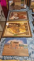 Three framed oil paintings laid flat on a table showing various subjects including pottery, a mountain lion, and adobe home.