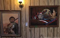View of two framed paintings hung on a wood-paneled wall: portrait of a buffalo hunter and a still life with harvest elements.