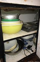 Yellow and white mixing bowls with green strainers on top, metal baking pans stacked on the right side, stored on an upper shelf.